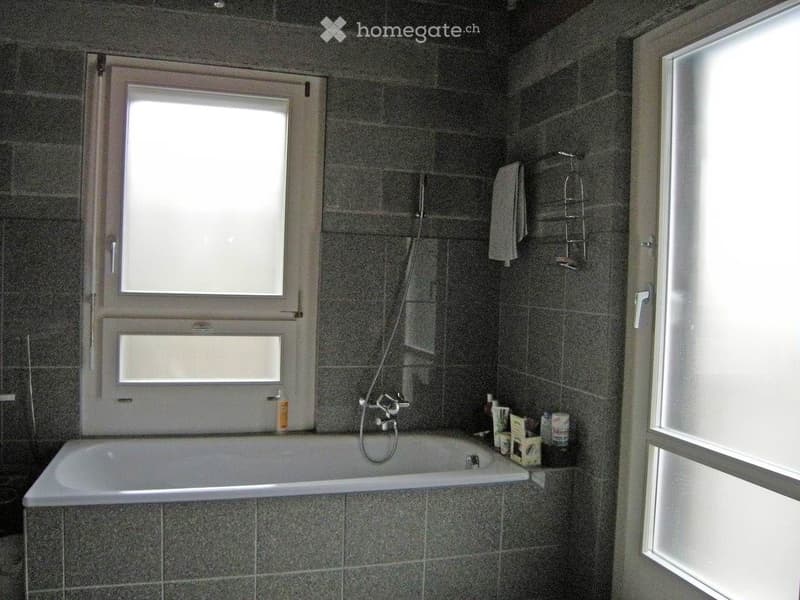 Modern bathroom with a bathtub, tiled walls, a window, and a towel rack with a towel. The bathtub has a faucet and a shower hose attached.