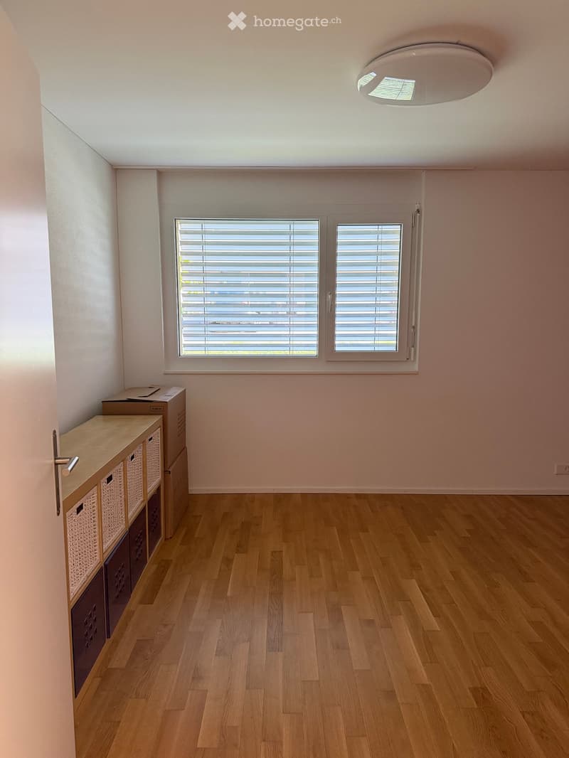 Empty room with light wooden flooring, white walls, and a large window with blinds. A small cabinet with drawers and wicker baskets is visible on the left side.