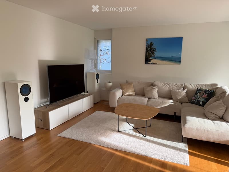 Modern living room with a large sectional sofa, wooden coffee table, floor lamp, speakers, and a large TV on a stand.