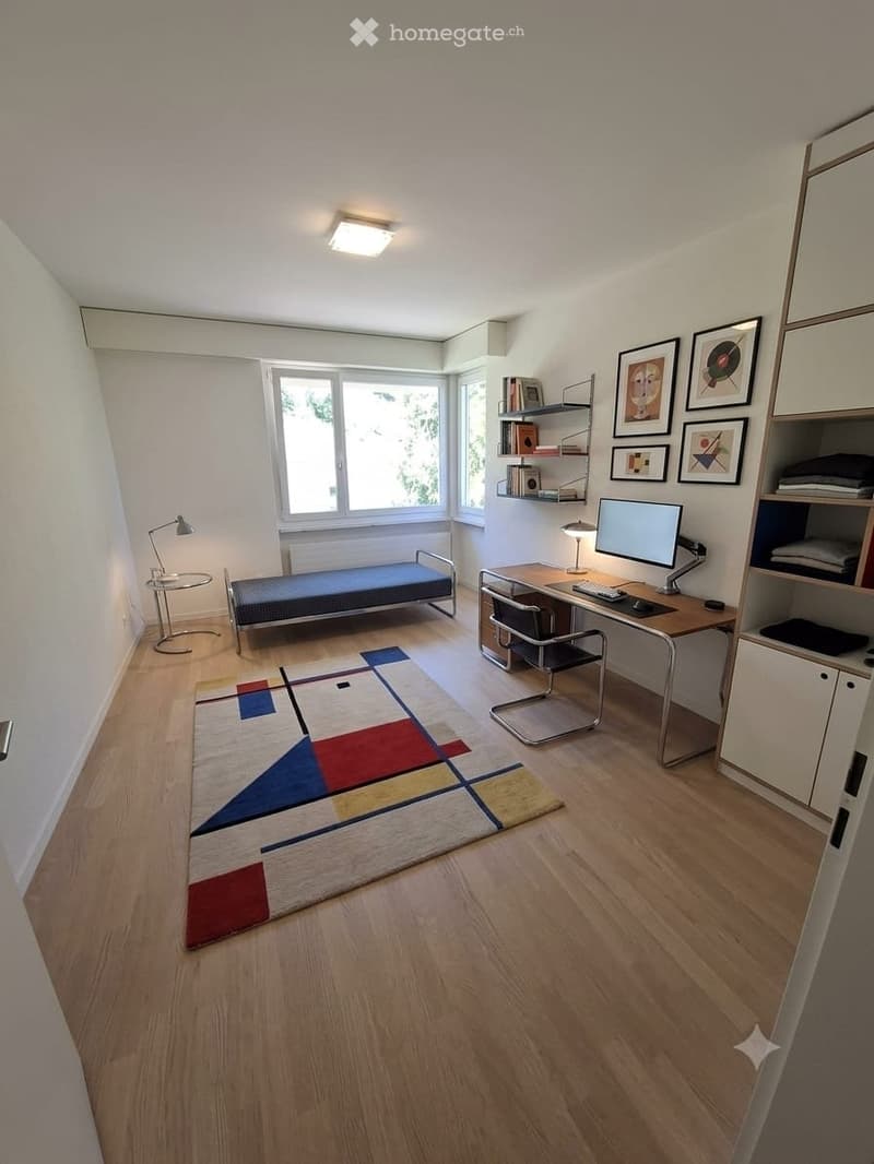 A modern, well-lit room with a desk, chair, screen, lamp, shelf, bed, small table, and picture frames on the wall.
