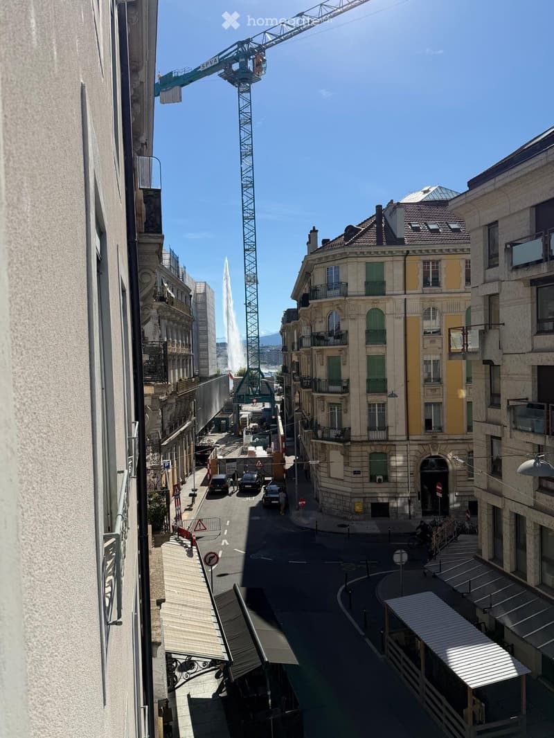 View from a balcony showing a city street with classic European architecture, a fountain, and a construction crane in the background.