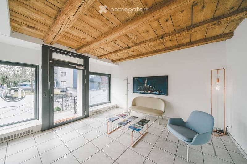 Modern living room with white walls, exposed wooden ceiling beams, tiled floor, large glass doors leading to a balcony, a beige sofa, a blue armchair, a glass coffee table with magazines, a floor lamp, and a television mounted on the wall.