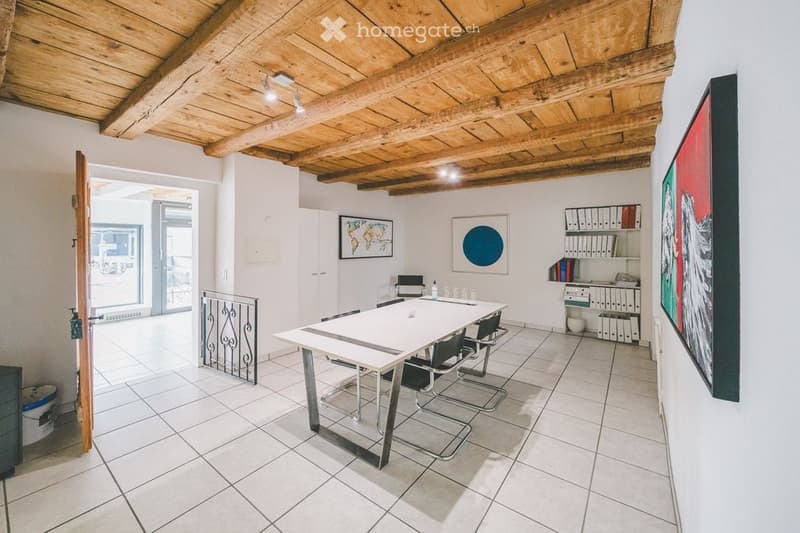 Bright, modern room with exposed wooden ceiling beams, white tiled floor, and large windows. Features a white conference table with four black chairs, a white shelving unit with binders and books, and several framed artworks on the walls.