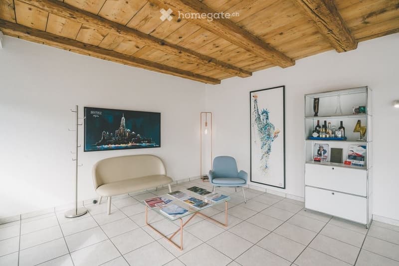 Modern living room with white tiled flooring, exposed wooden ceiling beams, a cream-colored sofa, a light blue armchair, a glass coffee table with magazines, a white cabinet with glass doors displaying bottles and decorative items, and two large framed ar