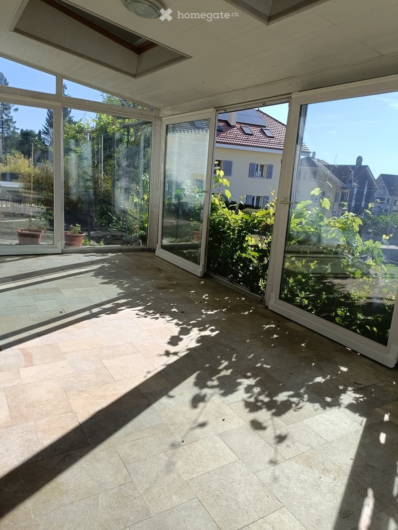 Bright, spacious balcony with large glass doors, tiled floor, and natural light. Shadows of plants are cast on the floor.