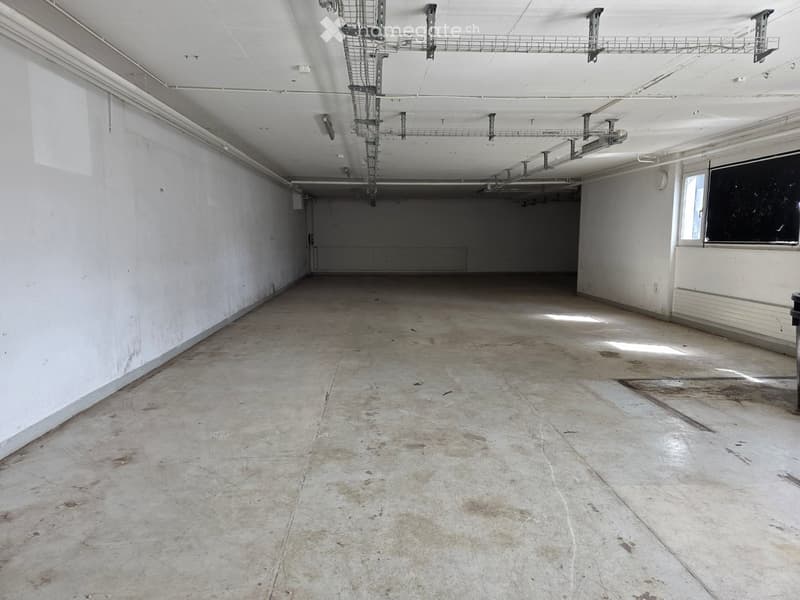 Empty room with bare concrete floor and white walls, exposed ceiling with metal shelving units, and a small window on the right side.