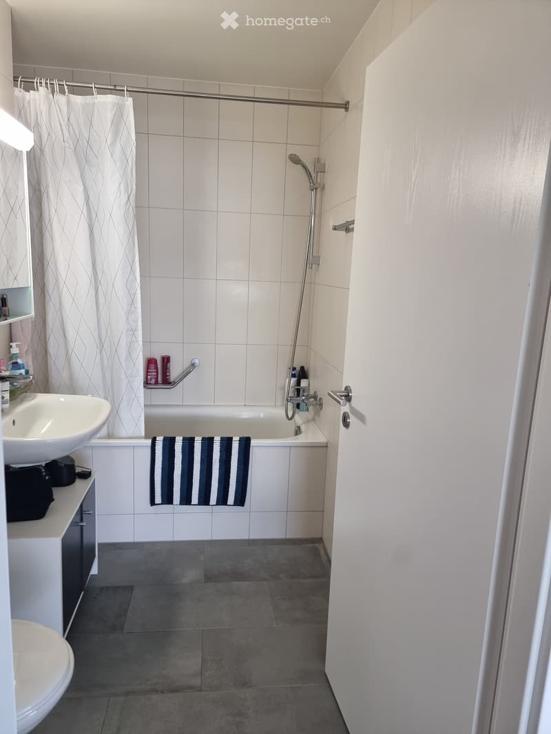 Modern bathroom with white tiles, bathtub, shower, sink, toilet, and towel. Features a shower curtain and a handrail for safety.