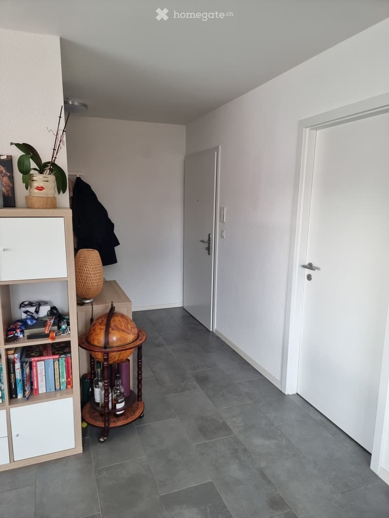 A hallway with a grey tiled floor, white walls, and a wooden shelf with books, a globe, a vase, and a coat hanging on the wall.