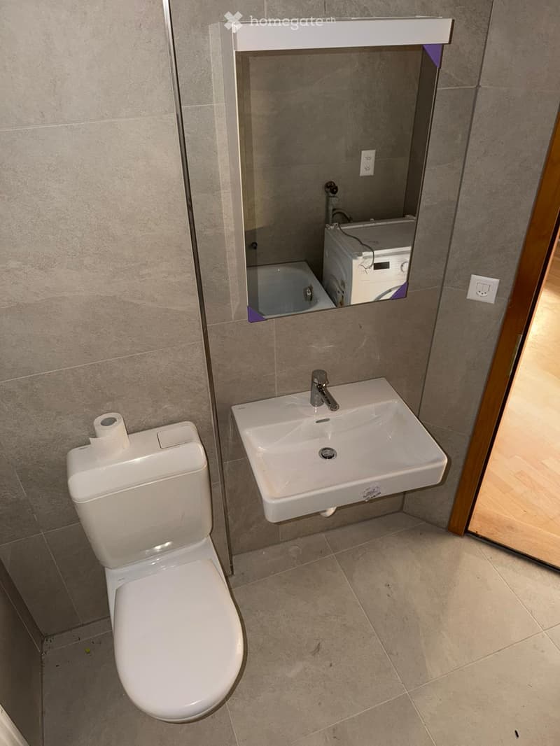 Modern bathroom with a white toilet, a wall-mounted sink with a chrome faucet, and a mirror above the sink. The walls are tiled in a neutral gray color, and the floor is tiled in a light beige color.