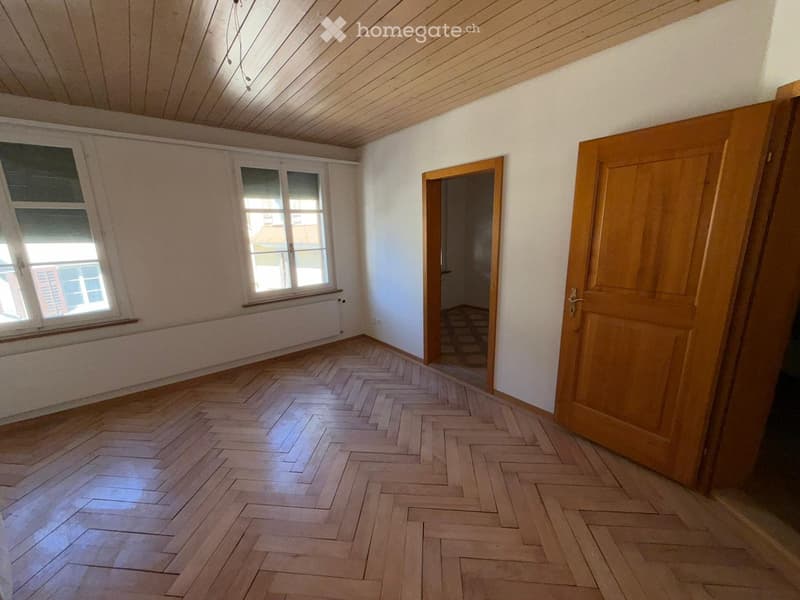 Empty room with wooden herringbone flooring, white walls, and wooden doors. Two windows with white frames and a radiator beneath one window. The ceiling is wooden with exposed beams.