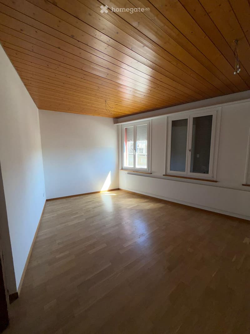 Empty room with wooden flooring, white walls, and wooden ceiling. Two windows with blinds allow natural light to enter.