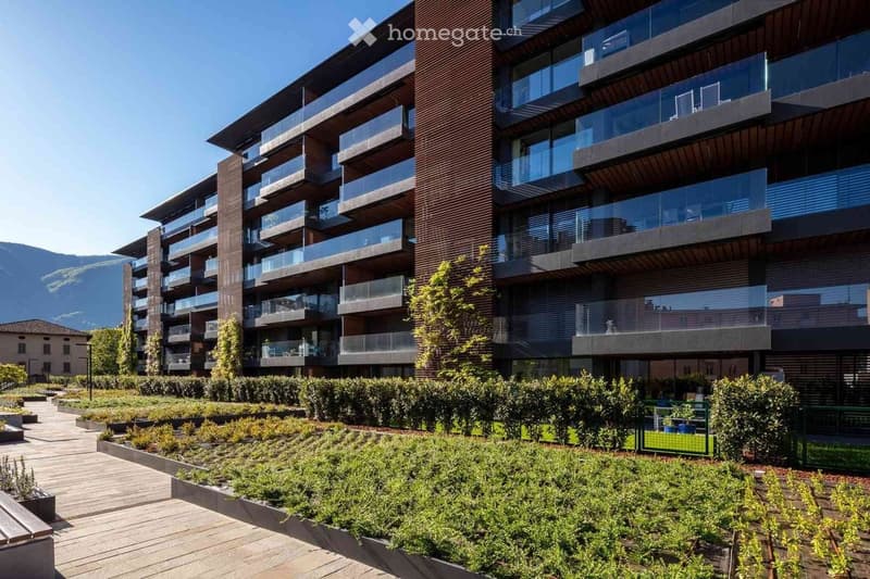 Modern multi-story residential building with balconies and large windows, surrounded by landscaped garden area with pathways and greenery.