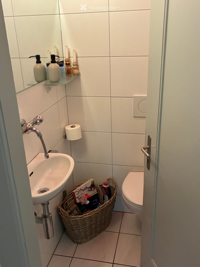 Modern bathroom with white tiled walls, a sink with a chrome faucet, a toilet, a wicker basket on the floor, and a glass shelf with candles and soap dispensers.