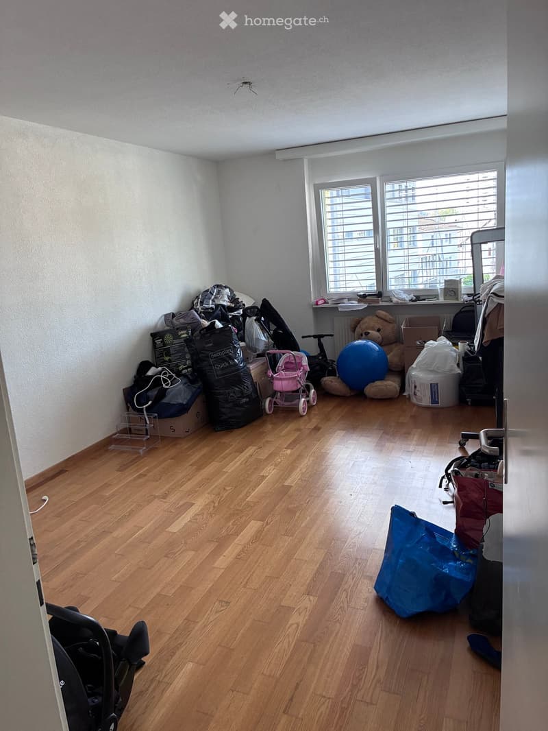 A room with wooden flooring and white walls, containing various items including a baby stroller, a large teddy bear, a blue exercise ball, a bicycle, and several bags and storage boxes.