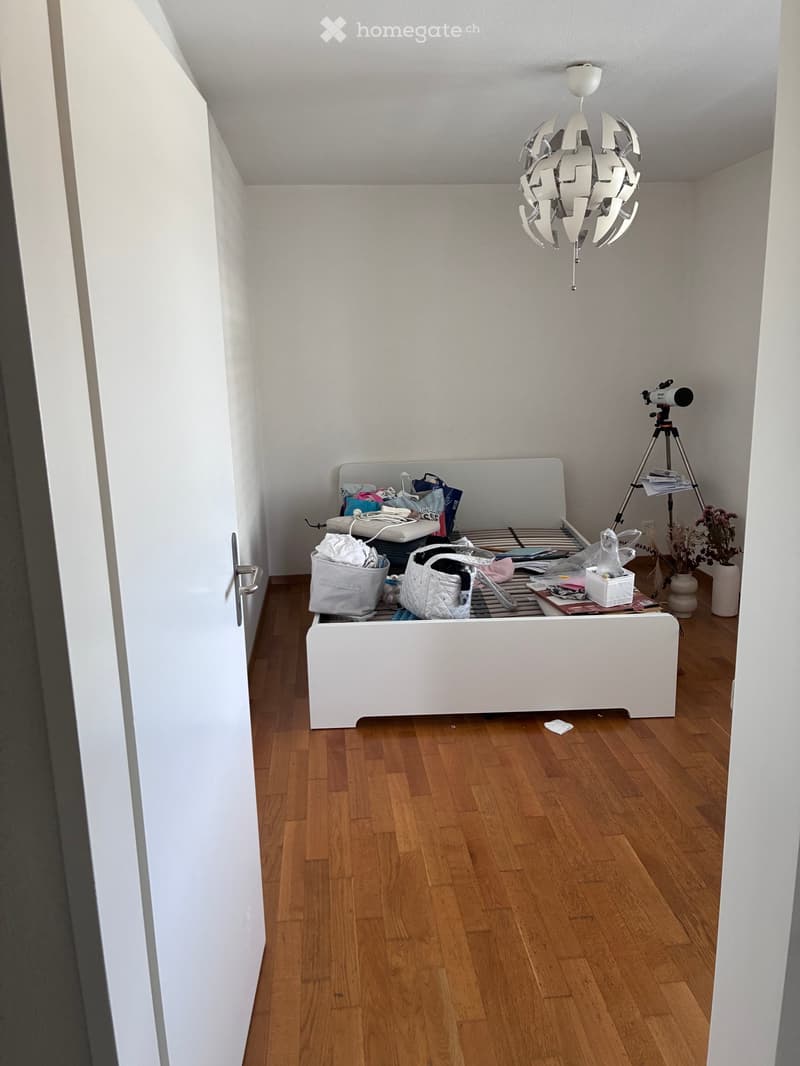 A small, modern bedroom with a white bed frame, wooden flooring, and a hanging geometric chandelier. A tripod with a camera is positioned near the bed, along with storage boxes, a handbag, and a potted plant in the corner.