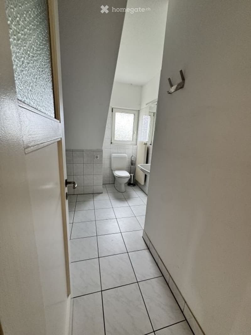 Small bathroom with white tiled floor and walls, toilet, sink, and a window for natural light.