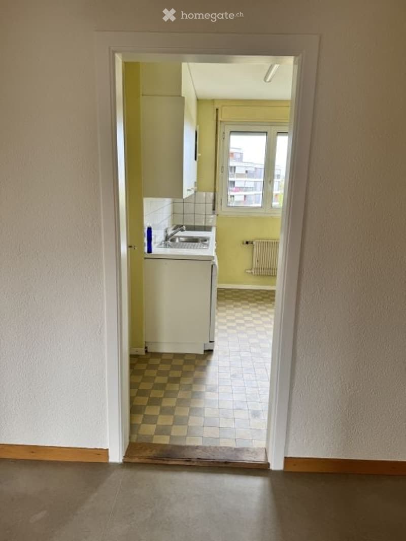 Empty room with a doorway leading to a small kitchen area. The kitchen has a sink, cabinets, and a window with a view of a building outside.