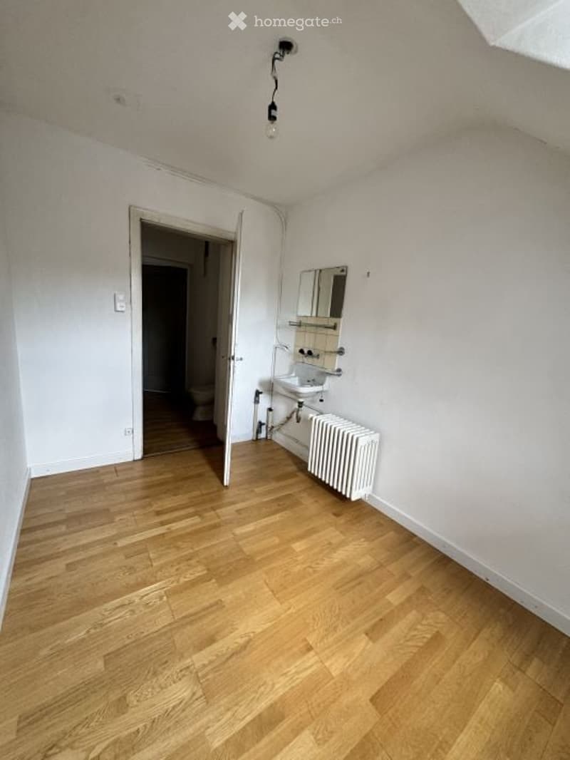 Empty room with wooden flooring, white walls, a radiator, a sink, and a mirror in the corner. The room is connected to a bathroom visible through an open doorway.