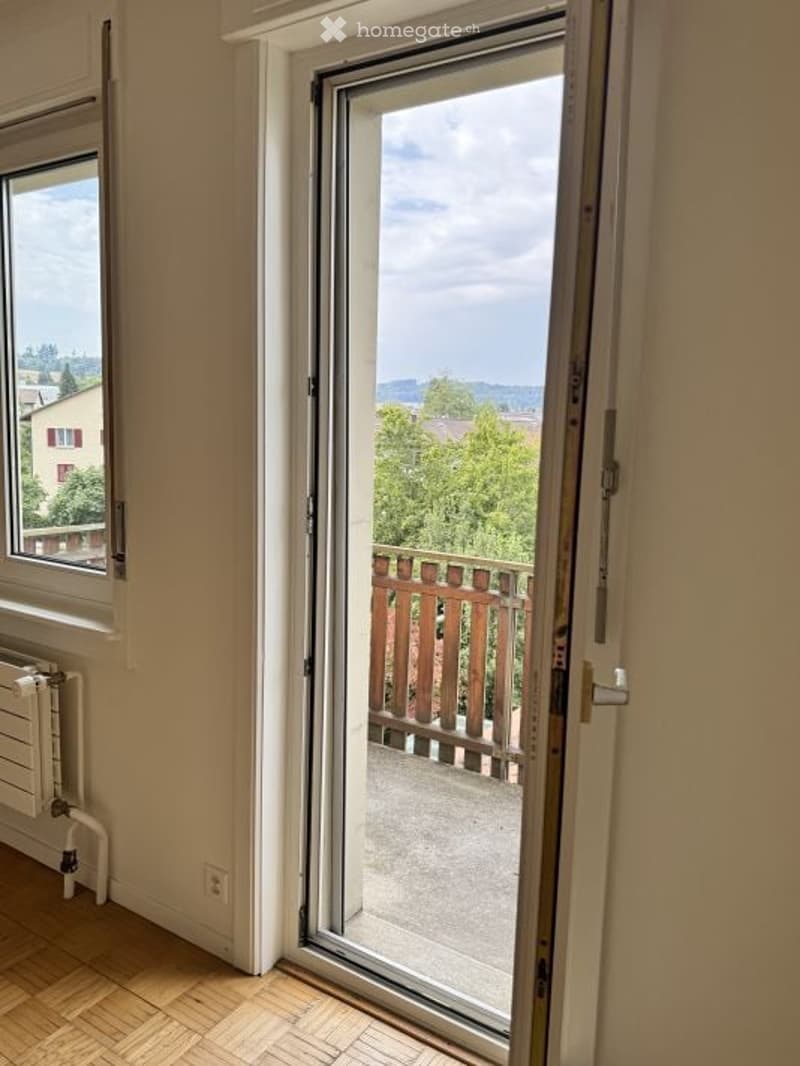 Empty room with wooden flooring, large window, and open sliding door leading to a balcony with wooden railing and scenic view.