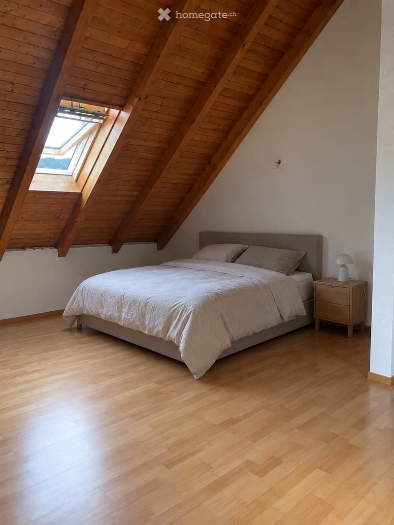 Bright, airy bedroom with wooden floors and slanted wooden ceiling. Features a large bed with white bedding, a nightstand with a lamp, and a skylight window bringing in natural light.