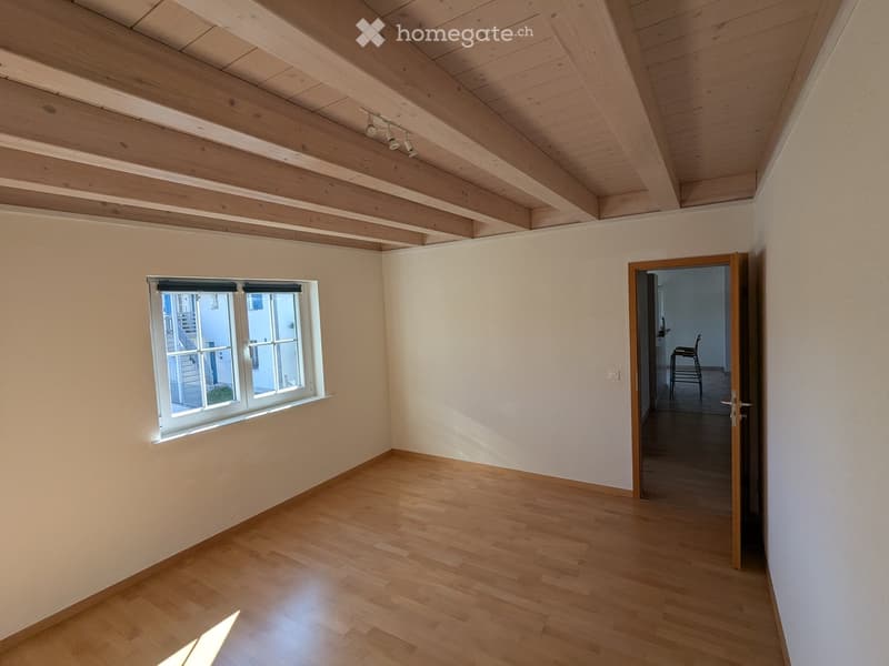 Empty room with wooden floor, white walls, exposed wooden ceiling beams, and a large window with blinds. A doorway leads to another room with a chair visible in the background.