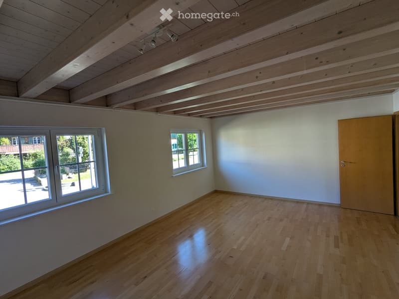 Empty room with wooden flooring, white walls, exposed wooden ceiling beams, and two windows, one of which is large and opens to the outside.