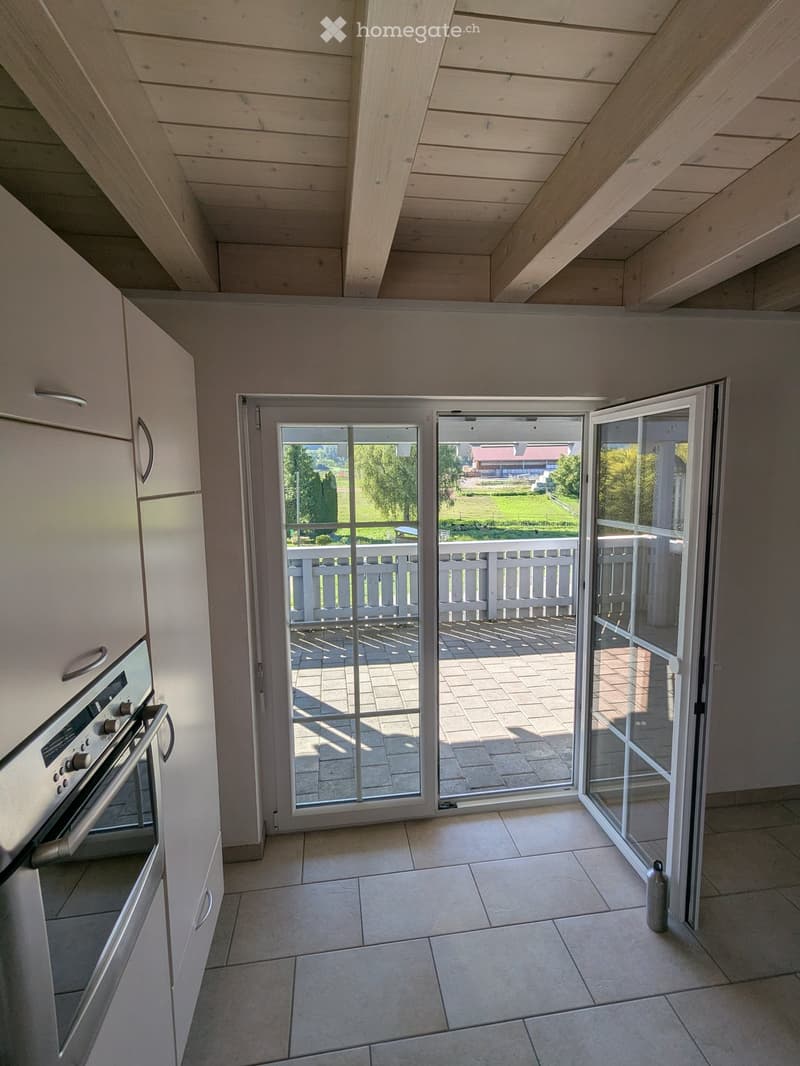 Modern kitchen with tiled floor, white cabinets, and an oven. Large sliding glass doors open to a patio with a white railing and a view of a green lawn.