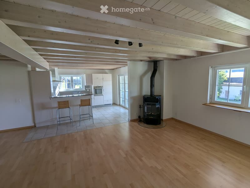 Open-plan living and dining area with wooden flooring, exposed wooden beams, a wood stove, and a small kitchen area with white cabinets and a bar counter.