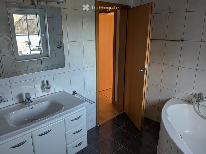 Modern bathroom with a white sink, bathtub, mirror, and cabinet. The room features tiled walls and a wooden door leading to another space.
