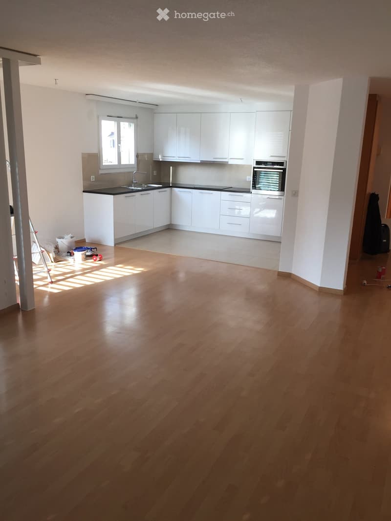 Empty kitchen with white cabinets, black countertops, and wooden flooring. The space is clean and well-lit with natural light from a window.