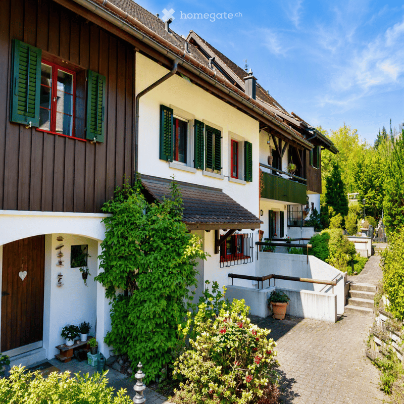Two-story house with white walls, red-framed windows, green shutters, and a tiled roof. The house features a wooden door, climbing plants, and a small garden area with potted plants and a pathway.