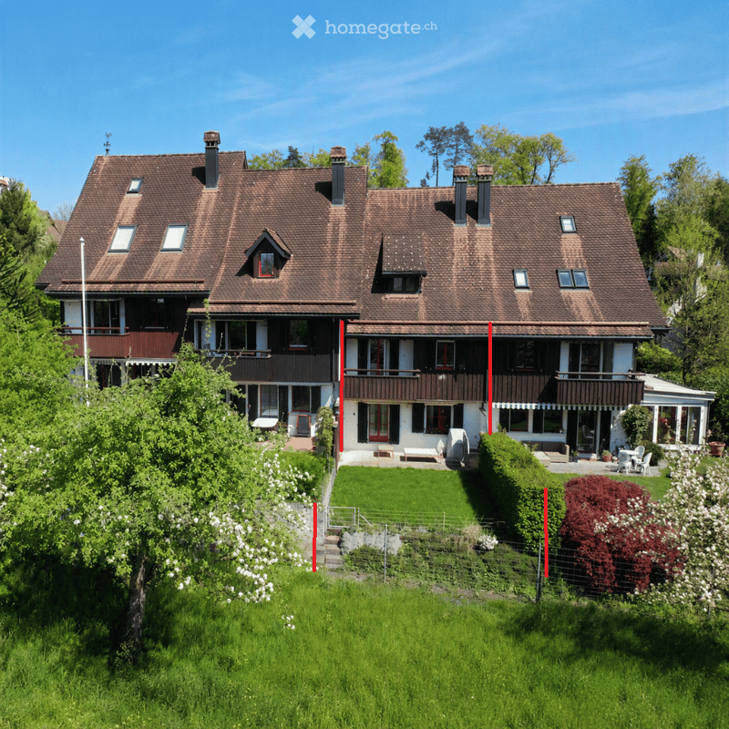 A two-story house with a brown tiled roof, multiple chimneys, and white walls. The house features large windows, balconies, and a well-maintained garden with green lawns, trees, and flowering shrubs.
