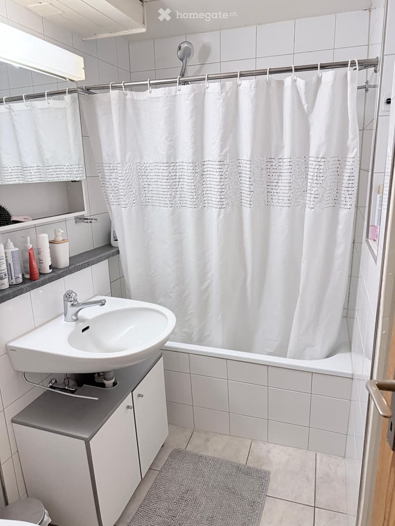 Modern bathroom with white tiles, a sink with a chrome faucet, a bathtub with a white shower curtain, a small cabinet, and a grey bathroom rug on the floor.