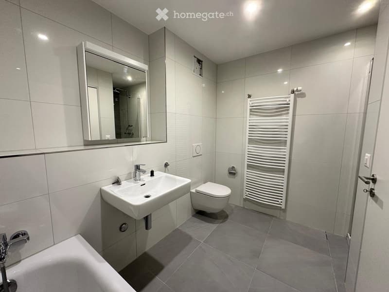 Modern bathroom with white tiles, a sink, toilet, mirror, and a white radiator. The space is clean and contemporary with grey flooring.