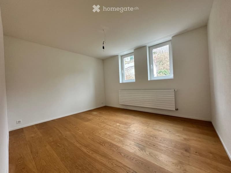 Empty room with wooden flooring, white walls, and a radiator. Two windows with white frames are visible, allowing natural light to enter.