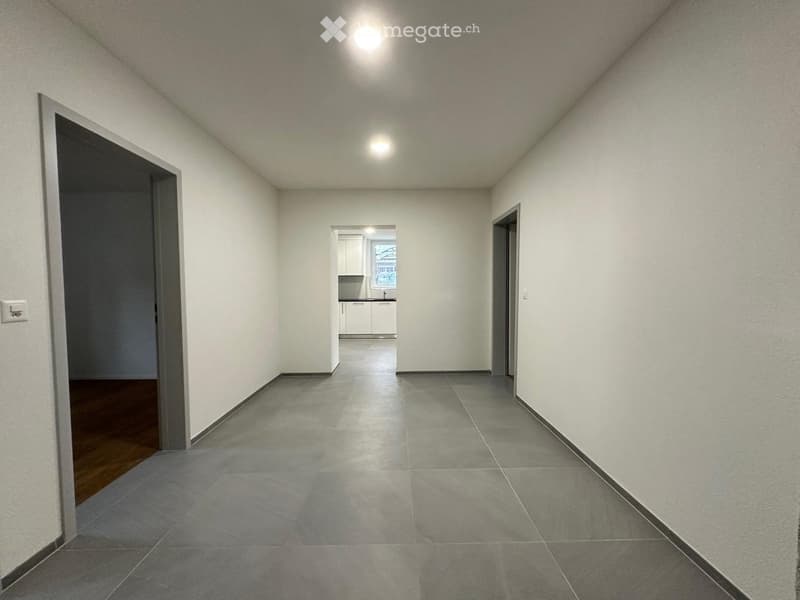 Empty hallway with grey tiled floor, white walls, and recessed lighting. Doors lead to adjacent rooms, including a kitchen visible in the background.