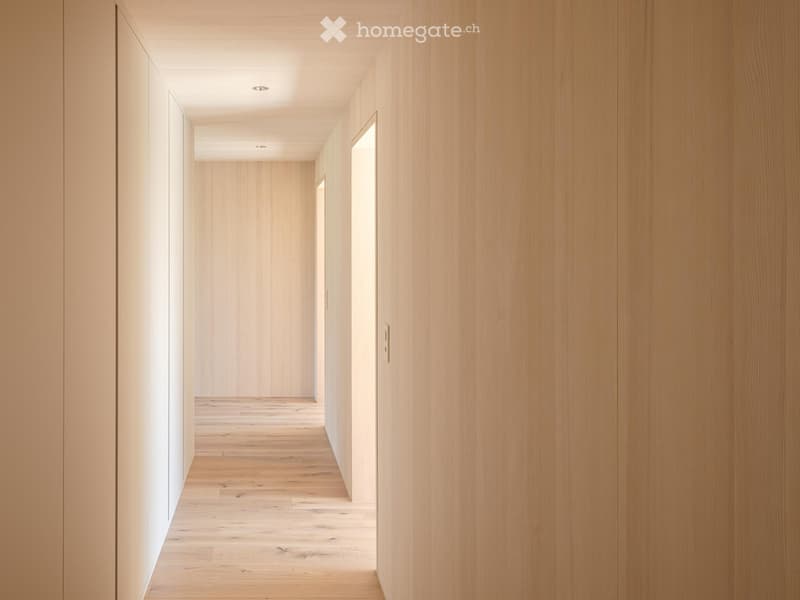 A long, narrow hallway with light wooden walls and flooring, featuring recessed ceiling lighting and a partially open door at the end.