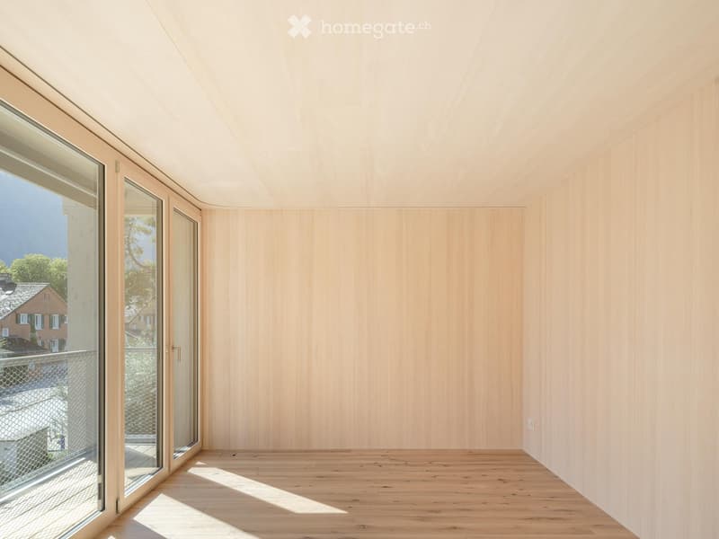 Empty room with light wooden walls and floor, large glass sliding doors leading to a balcony, and natural light streaming in.