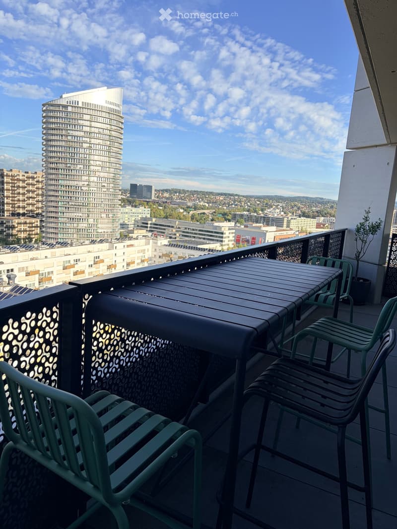 Balcony with a table and chairs, overlooking a cityscape with modern buildings and a clear blue sky.