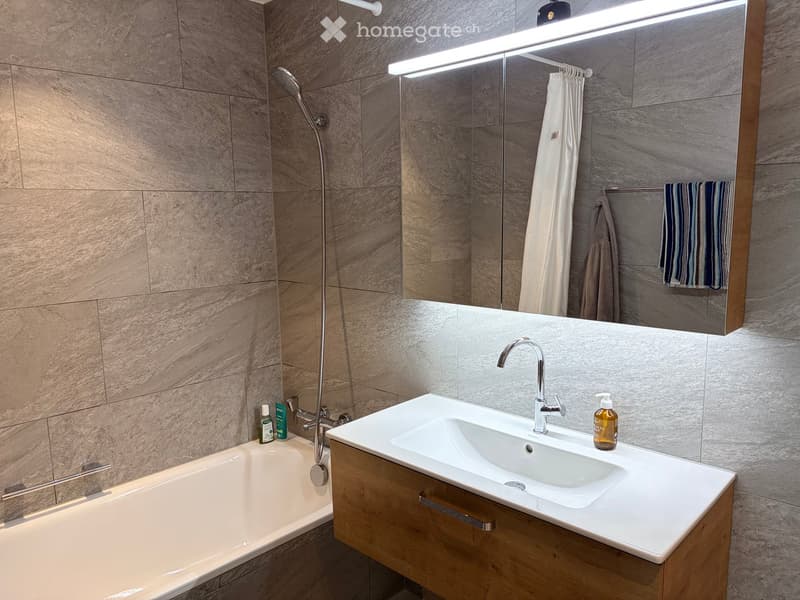 Modern bathroom with a bathtub, sink, and showerhead. Features a large mirror with built-in lighting, a wooden vanity, and tiled walls.