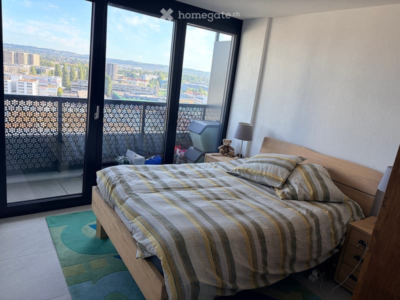 Cozy bedroom with a wooden bed, striped bedding, a nightstand with a lamp, a teddy bear, and a view of the city from large windows.