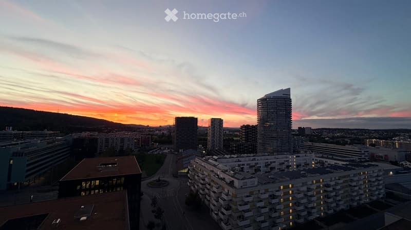 A panoramic view of a cityscape at sunset, featuring modern high-rise buildings, residential complexes, and a winding road, with a distant mountain range and colorful sky.