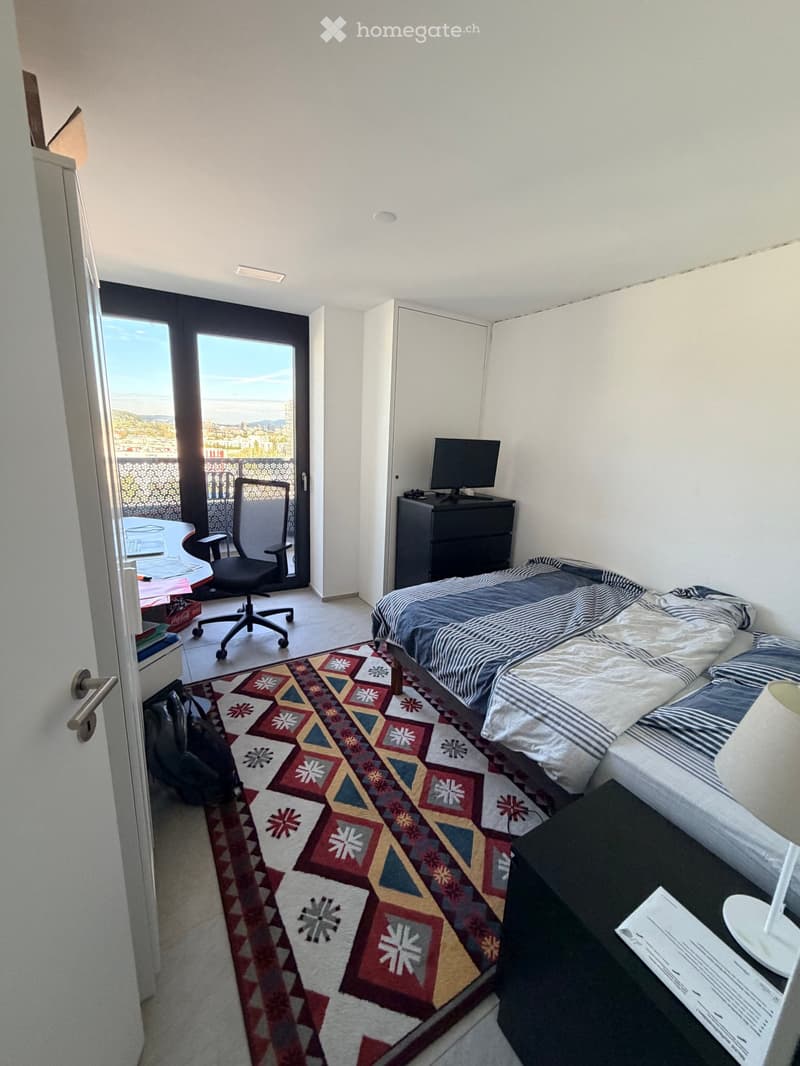 Cozy bedroom with a bed, chair, table, laptop, nightstand, and lamp. Features a colorful geometric rug and a view of the city from a balcony.