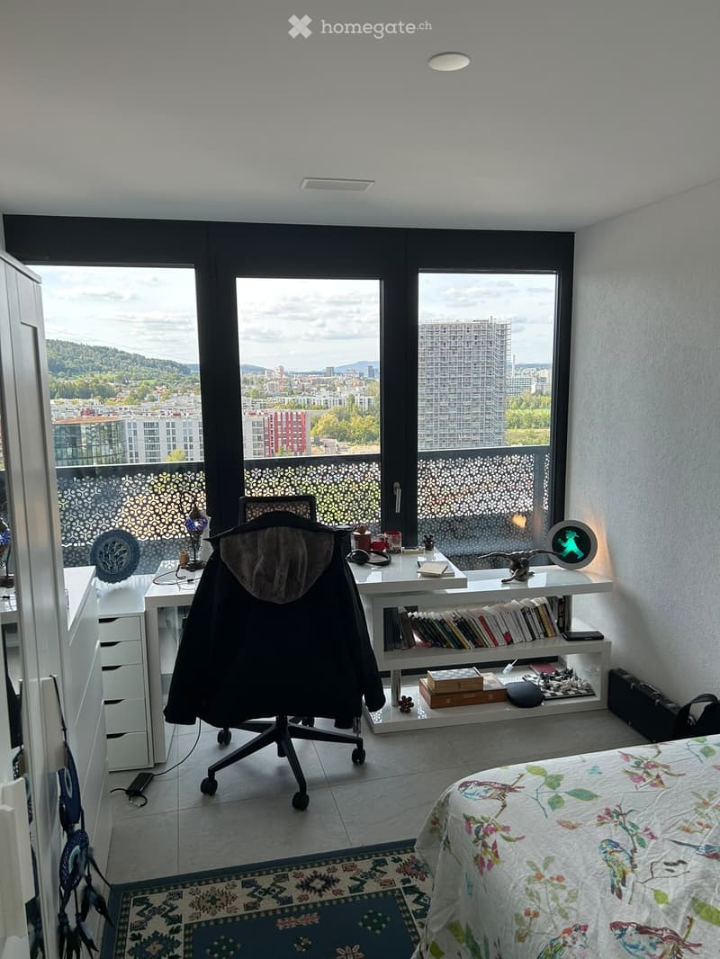 Modern bedroom with a bed, desk, chair, shelf, and large windows offering a city view.
