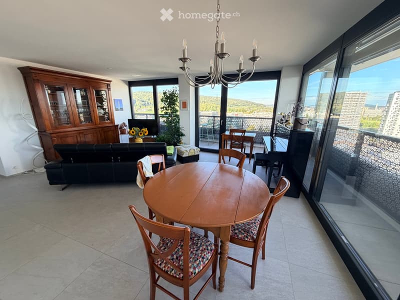 Bright living and dining area with large windows offering a scenic view, featuring a sofa, dining table with chairs, television, and a wooden cabinet.
