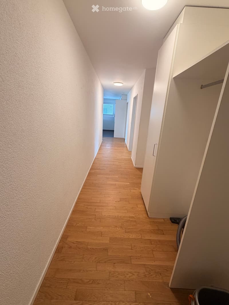 A narrow hallway with wooden flooring, white walls, and recessed lighting. The hallway leads to a room with a window and a door at the end.