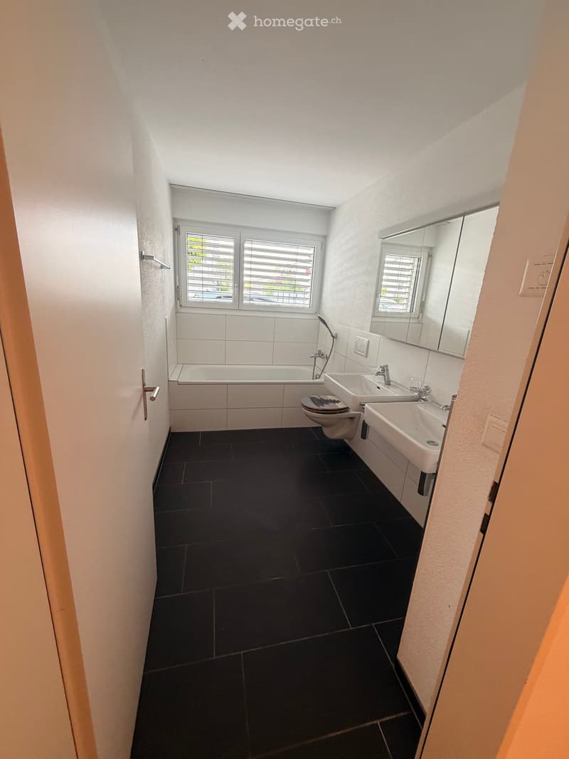 Modern bathroom with dark tiled floor, white tiled walls, bathtub, two sinks, toilet, and a large mirror above the sinks.
