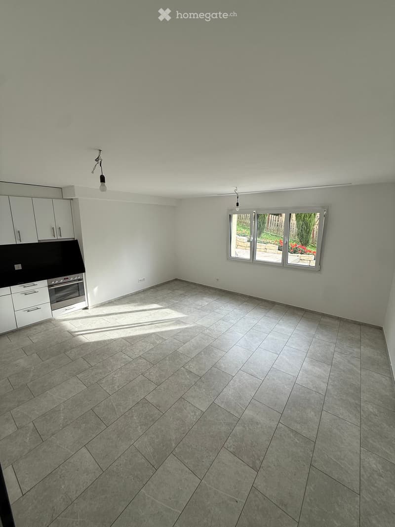 Empty room with gray tiled floor, white kitchen cabinets, black countertop, stainless steel oven, glass window with sliding doors and view of garden with flowers.