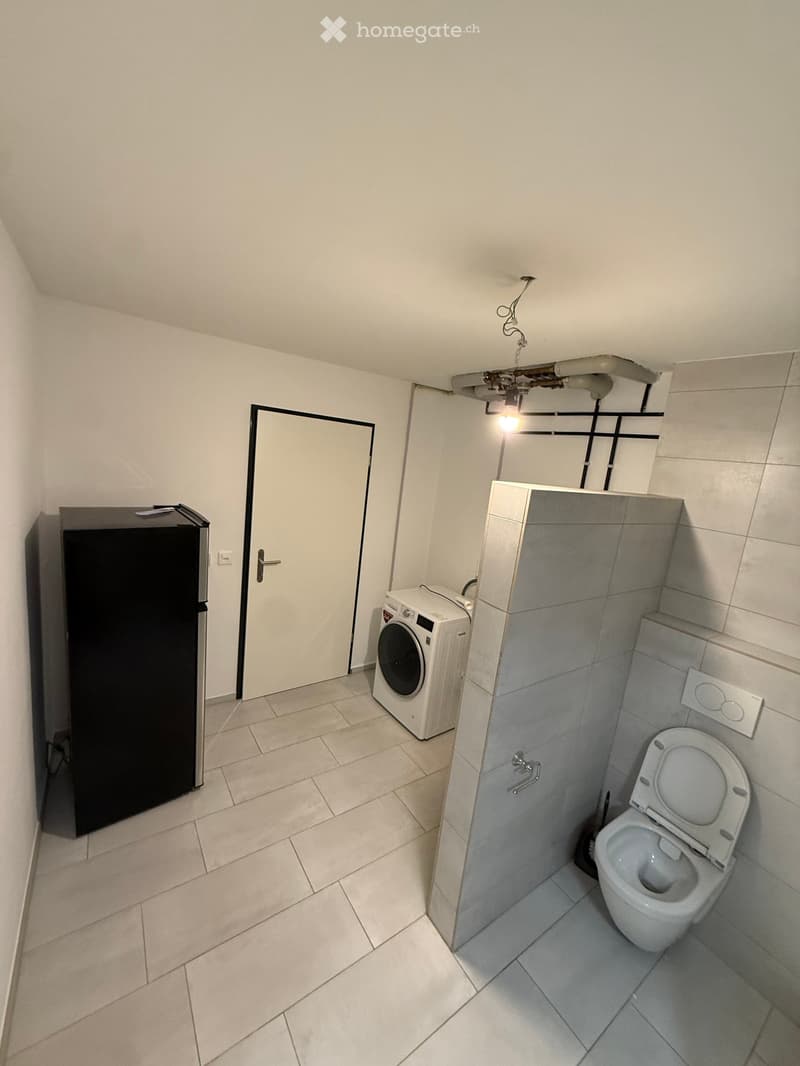 White tiled floor, white walls, a washing machine, a refrigerator, a white toilet, a shower stall, an open door