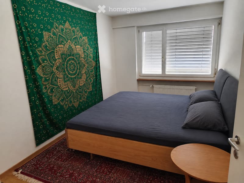 Cozy bedroom with a wooden bed, dark blue bedding, a small wooden table, and a green mandala-patterned curtain on the wall.